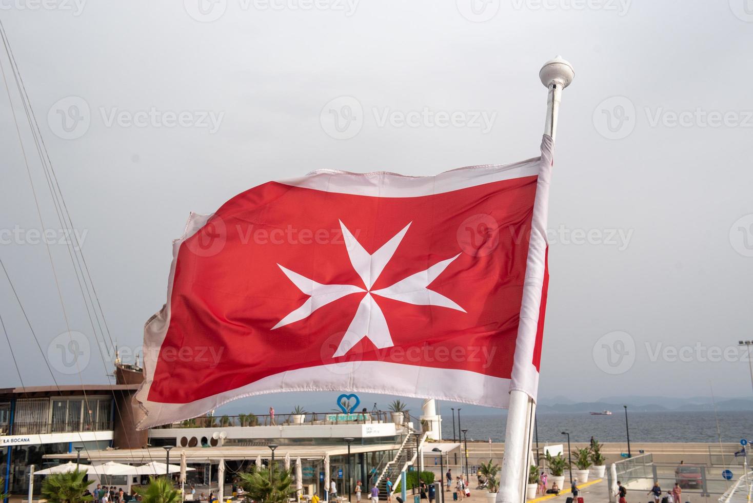 Maltese flag on a luxury boat anchored in the marina of Greece. Old Maltese flag, white cross on red background waving at stern of yacht. Blue sky background. photo
