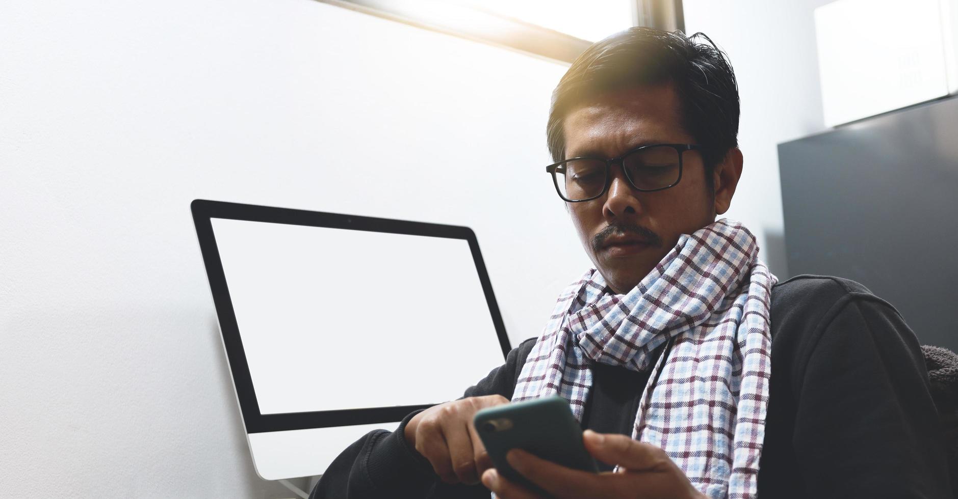 An Asian man uses a phone while working on a desk with a computer. photo