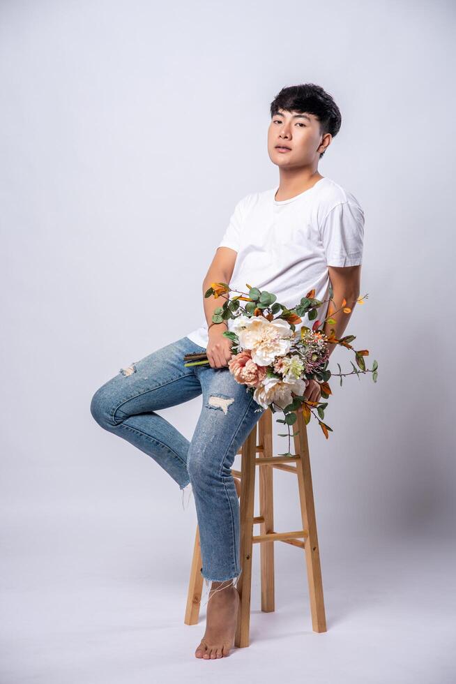 A young man in a white t-shirt sits on a high chair and holds flowers. photo