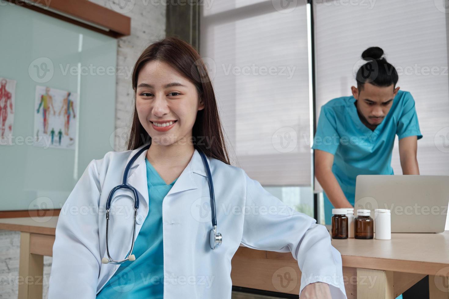 Portrait of beautiful female doctor of Asian ethnicity in uniform with stethoscope. Smile and looking at camera in a hospital clinic, male partner working behind her, two professional persons. photo