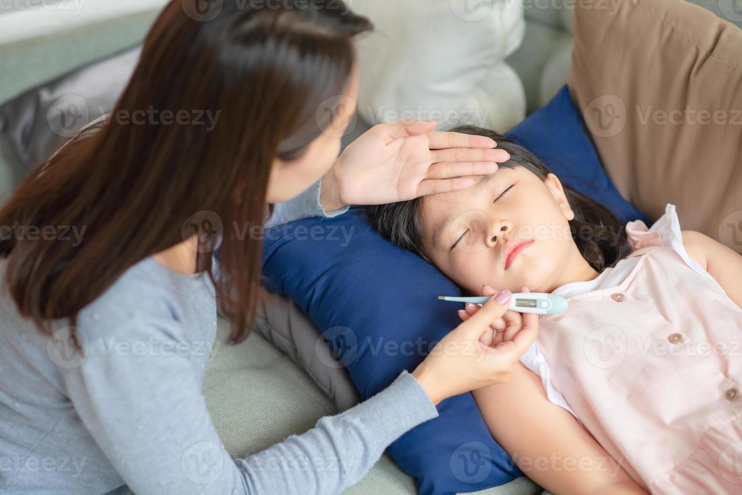 Asian mother checking her kid body temperature by thermometer which got fever and illness at home. photo