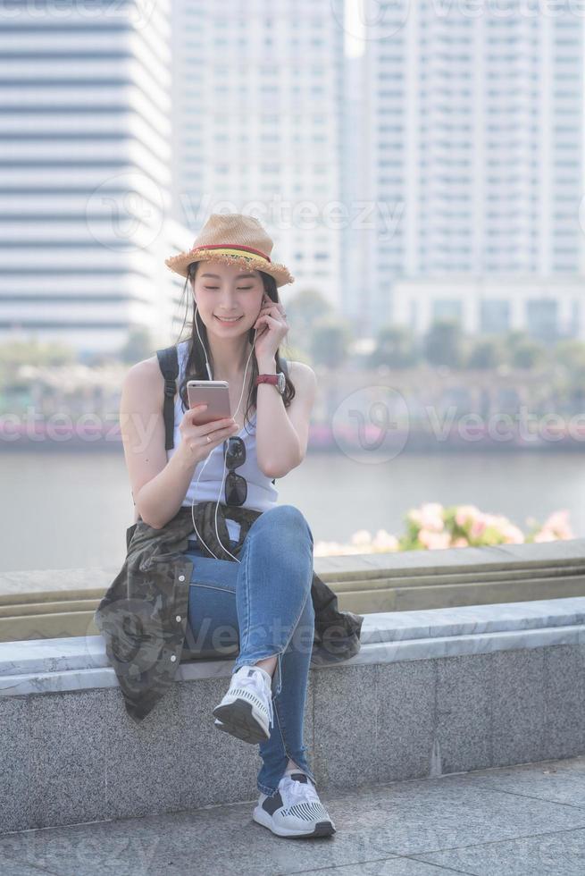 Beautiful asian solo tourist woman relaxing and enjoying listening the music on a smartphone in urban city downtown. Vacation travel in summer. photo