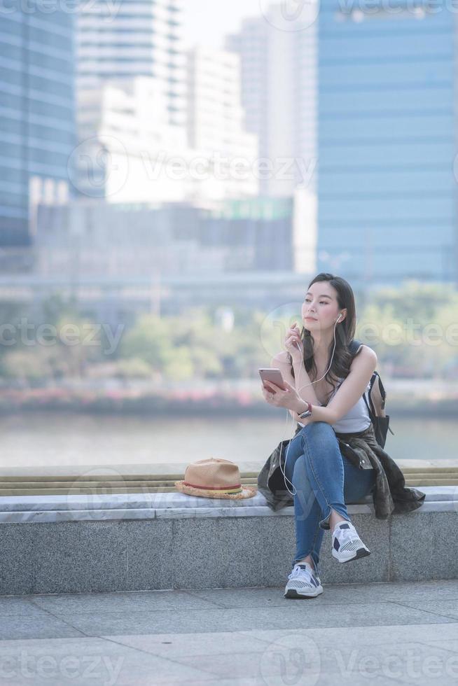 Hermosa mujer asiática turista en solitario relajándose y disfrutando escuchando música en un teléfono inteligente en el centro urbano de la ciudad. viajes de vacaciones en verano. foto