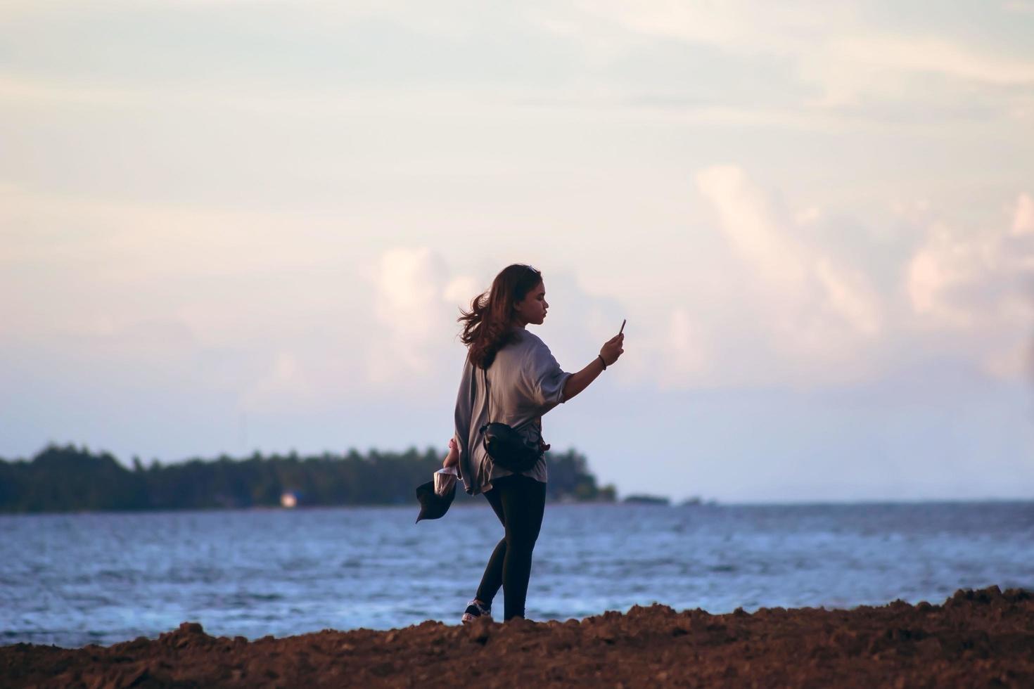 sorong, papua occidental, indonesia, 19 de noviembre de 2021. una niña sola en la playa tomando un autorretrato con un teléfono inteligente. tiempo personal. foto