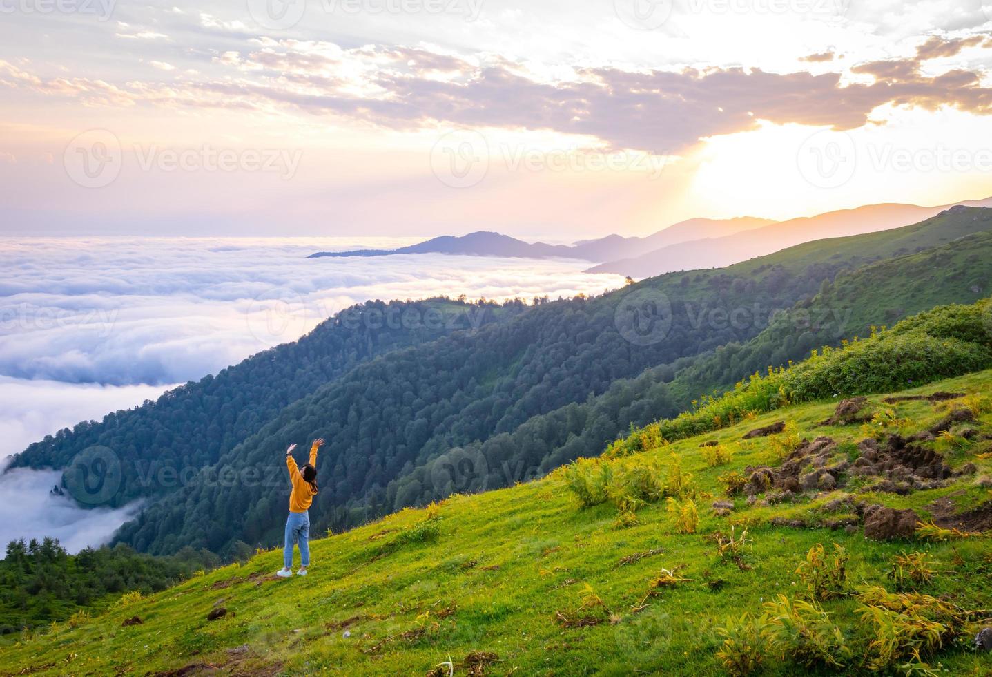 Female person out of joy looks to camera with spreaded hands and scenic landscape of Gomismta mountain above the clouds with sunrise in backgorun photo