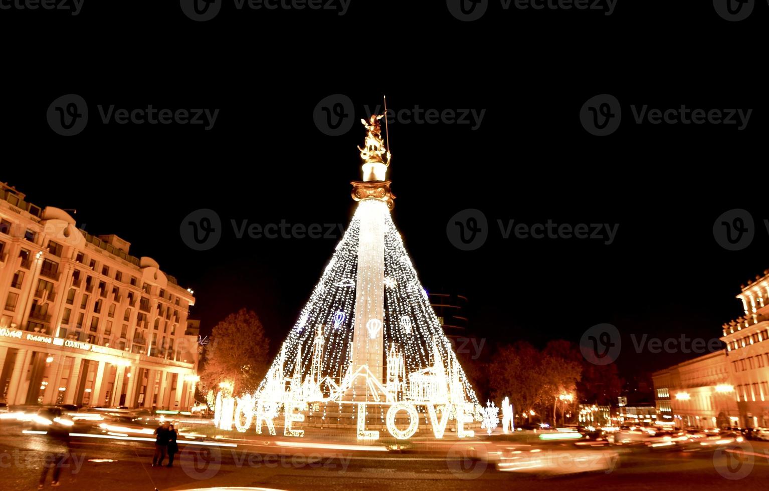 Cars pass on roundabout leaving light trails with decoration lights on statue in freedom square while tourist taking photographs photo