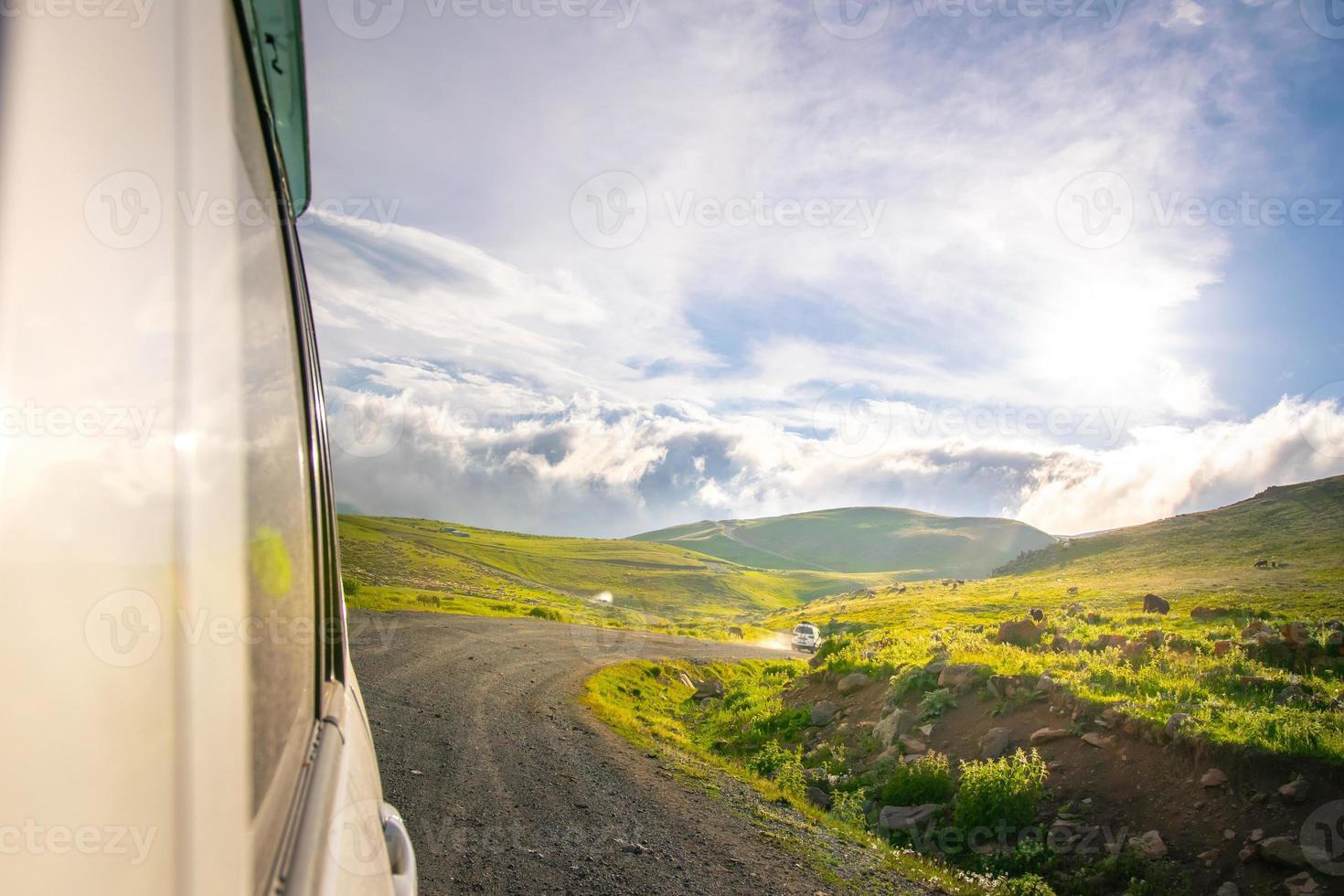 White car drives on caucasian green wilderness with sky and meadow view. Copy paste road trip background photo