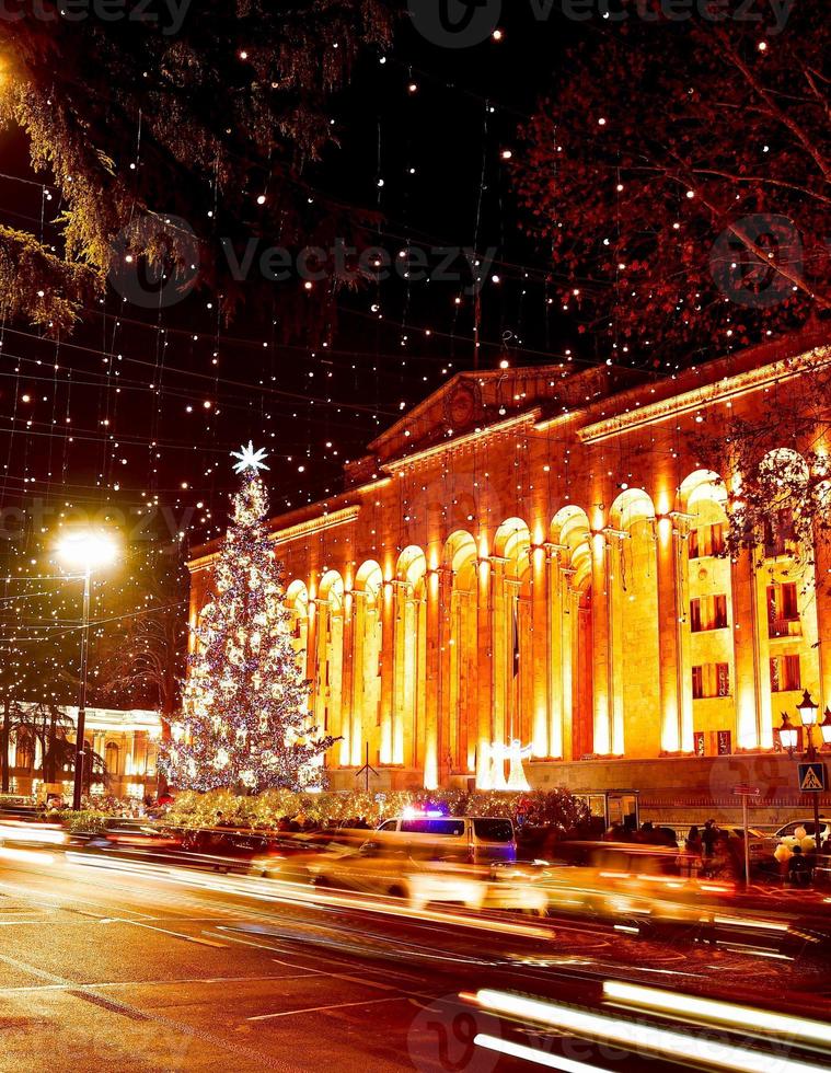 Coches en la carretera en el tráfico en horas punta por la noche en la avenida Rustaveli en la ciudad capital de Tbilisi en Georgia en Navidad con el árbol de Navidad por el parlamento en el fondo foto