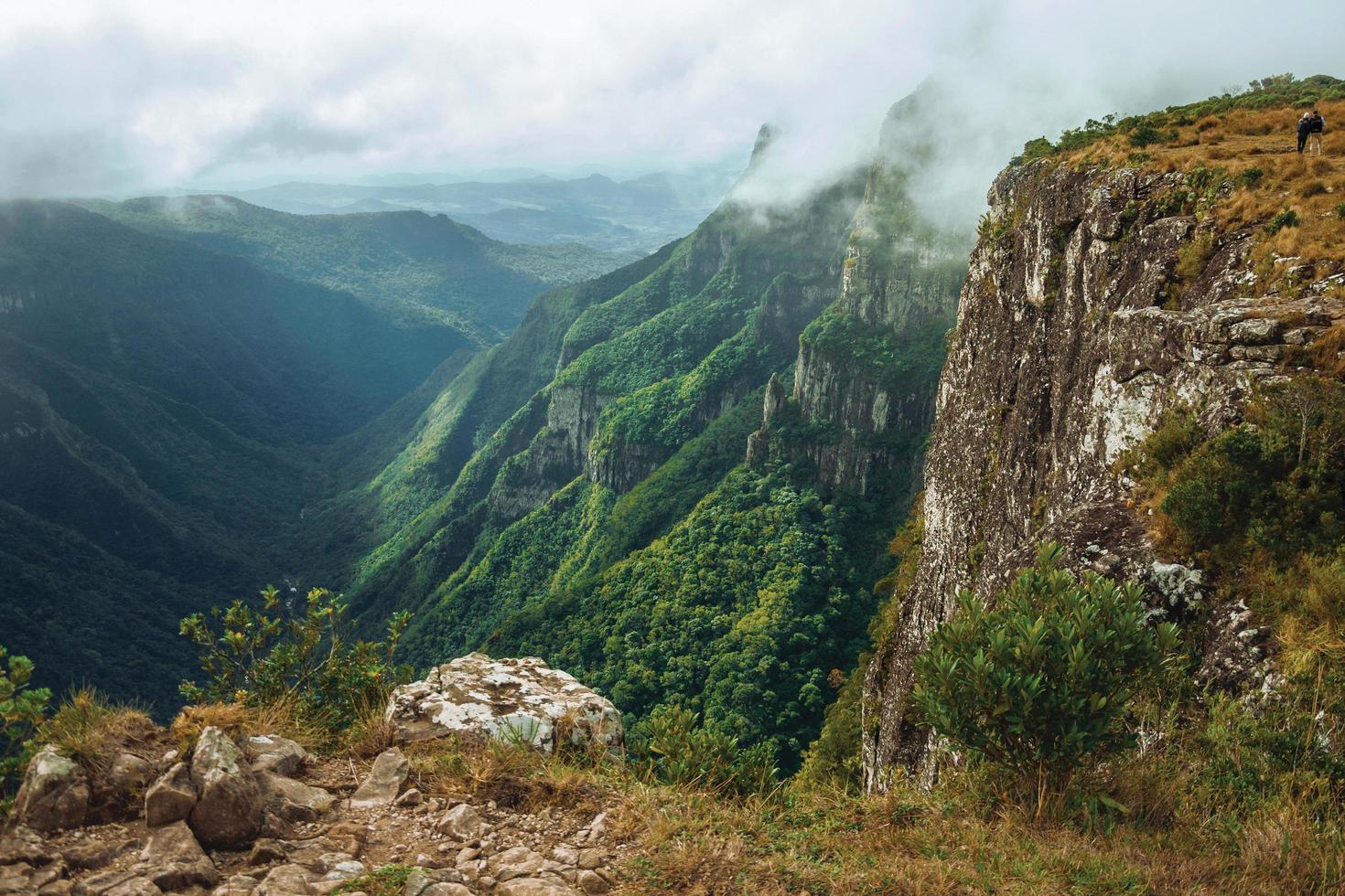 People on steep rocky cliff at Fortaleza Canyon covered by forest and mist coming up of the valley near Cambara do Sul. A small rural town in southern Brazil with amazing natural tourist attractions. photo