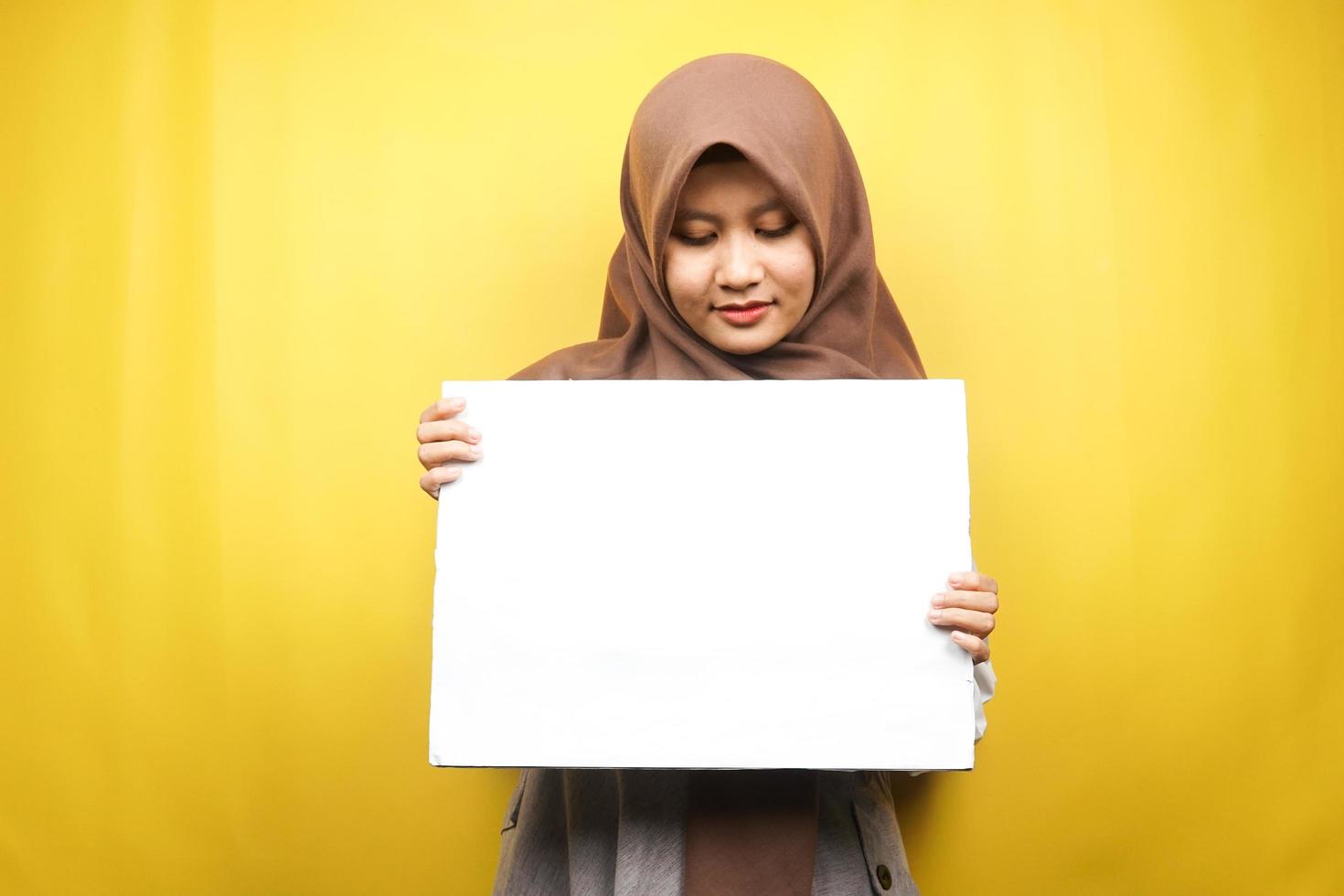 Bastante joven mujer musulmana alegre sosteniendo pancarta vacía en blanco, cartel, tablero blanco, tablero de letrero en blanco, tablero de anuncio blanco, presentando algo en el espacio de la copia, promoción foto