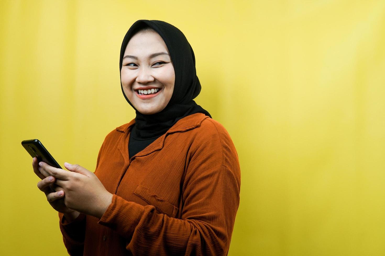 Hermosa joven musulmana asiática con manos sosteniendo teléfono inteligente, sonriendo confiada, entusiasta y alegre, ver mirando el espacio vacío presentando algo, aislado, concepto publicitario foto