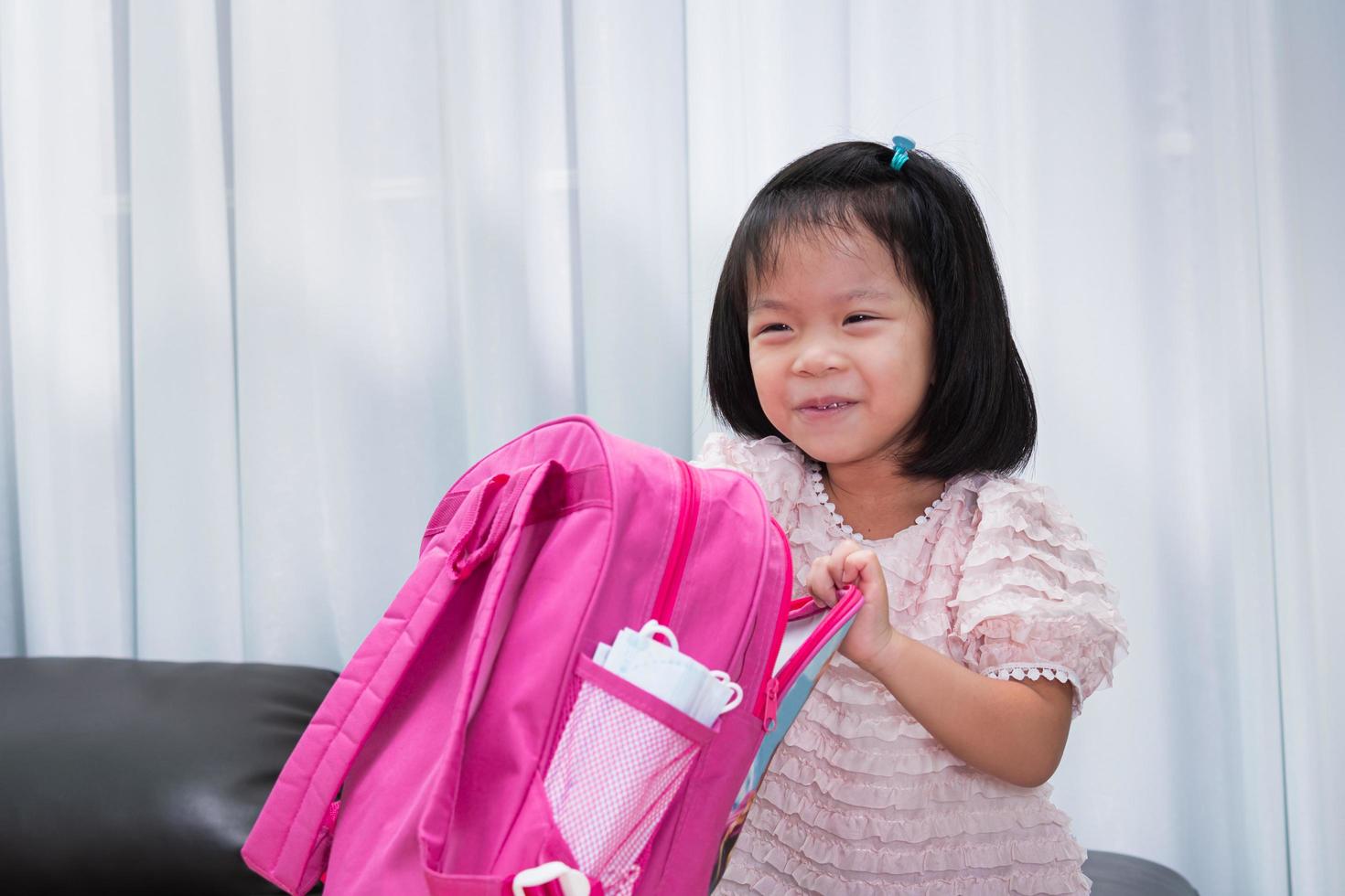 Kindergarten girl enjoyed preparing her pink school bags to go back to school. Medical mask is on side of bag. Empty space to enter text. Kid aged 4 years old. photo
