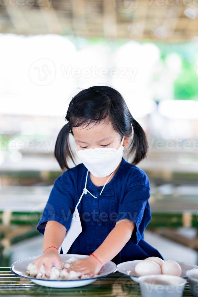 Kindergarten students are having fun mashing up preservative ingredients in zinc plate. Kid girl learn outside classroom. Child wear white surgical face mask. Children aged 4 year old. Vertical image. photo