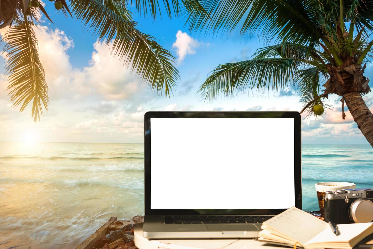 Mockup of laptop computer with empty screen with camera,notebook,coffee cup on table at landscape early sunrise over blue the sea background,working on the beach,Freelance work and holiday traveler. photo