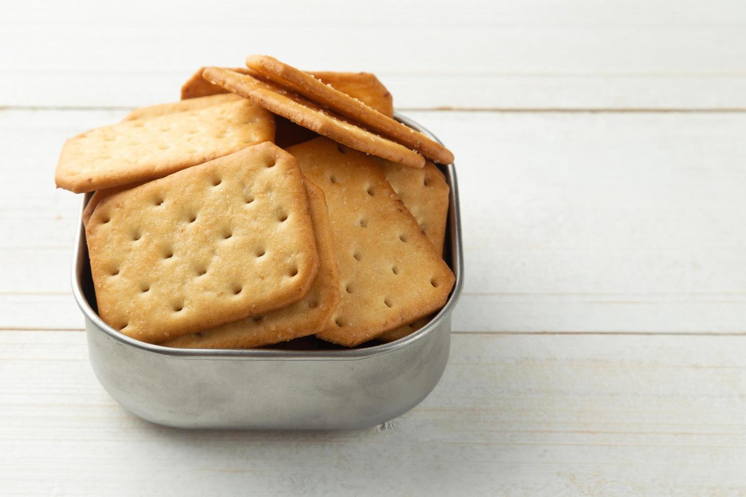 Galletas cracker en un recipiente de acero inoxidable sobre fondo de mesa de madera blanca foto