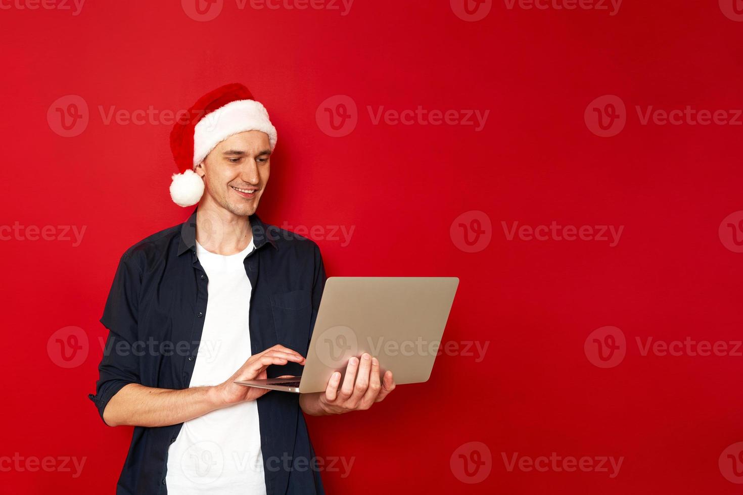 Laughing young man in casual blue shirt christmas hat posing isolated on red background studio portrait. People sincere emotions lifestyle concept. Mock up copy space. Working on laptop pc computer photo