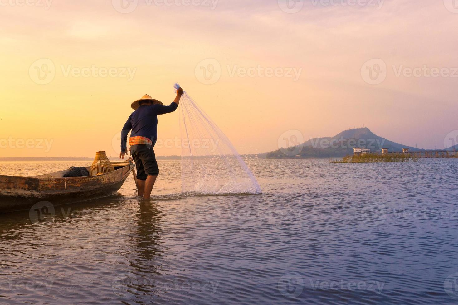 Asian fisherman with wooden boat throwing a net for catching freshwater fish in nature river in the early during sunrise time photo