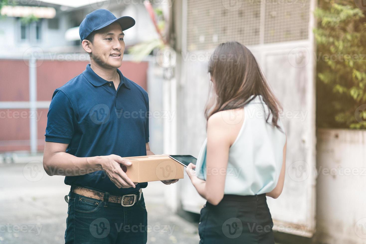 Handsome young asian delivery man smiling and holding a cardboard box while waiting beautiful young asian woman putting signature in smart tablet for confirm receive. photo