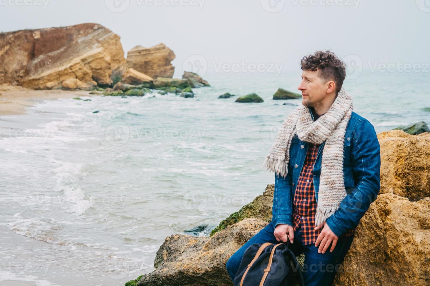 Traveler man with backpack standing on sandy beach in the middle of rocks against sea background photo