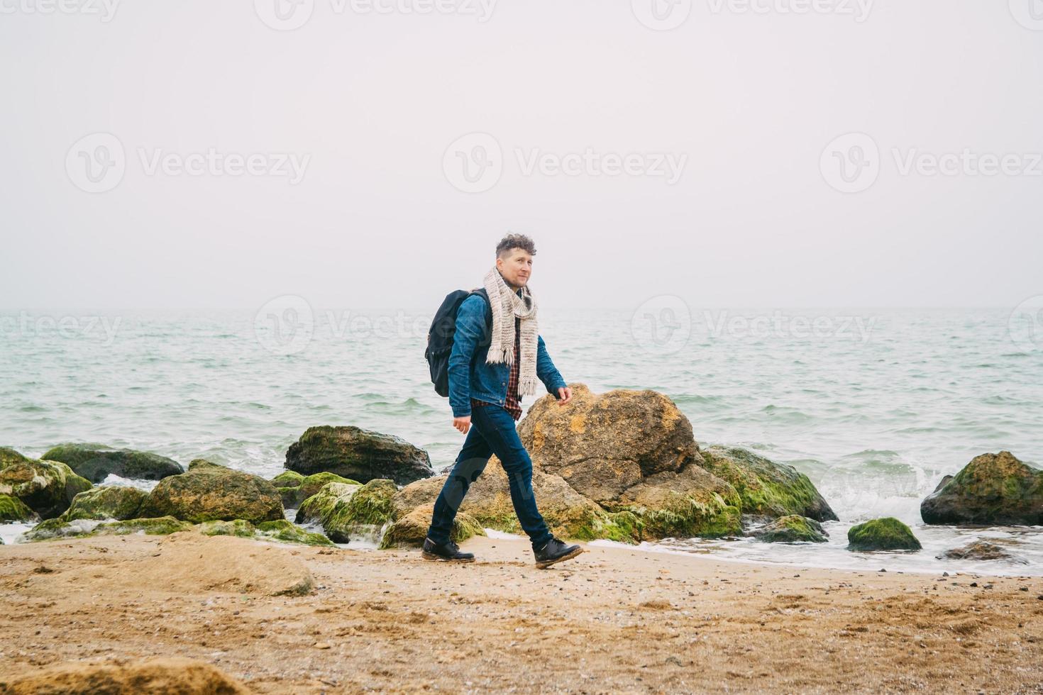 Traveler man with backpack standing on sandy beach in middle of rocks against sea background photo