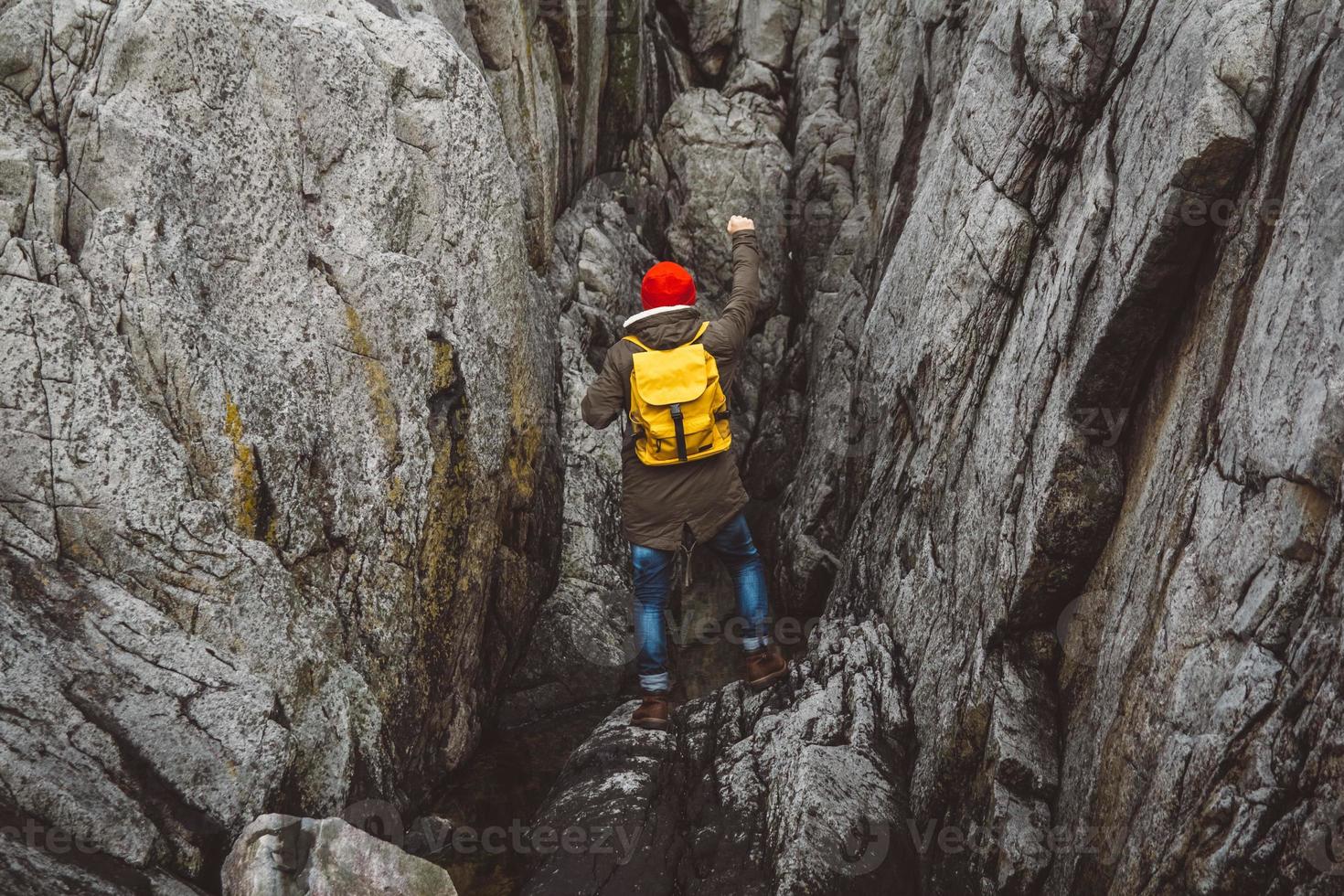 Traveler man with a yellow backpack standing on background of the rocks with a raised arm photo