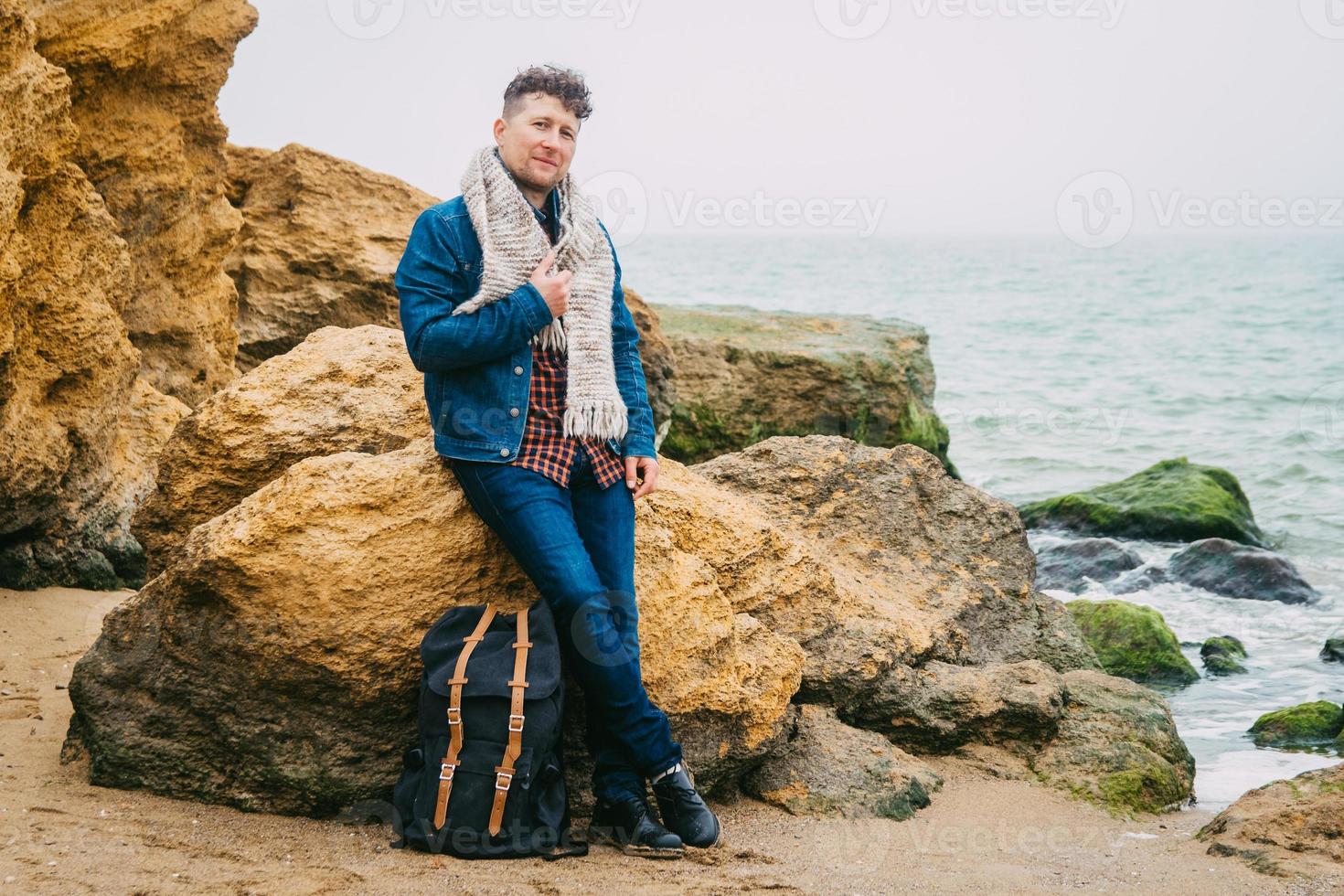 Hombre viajero con mochila de pie en la playa de arena en medio de las rocas contra el fondo del mar foto