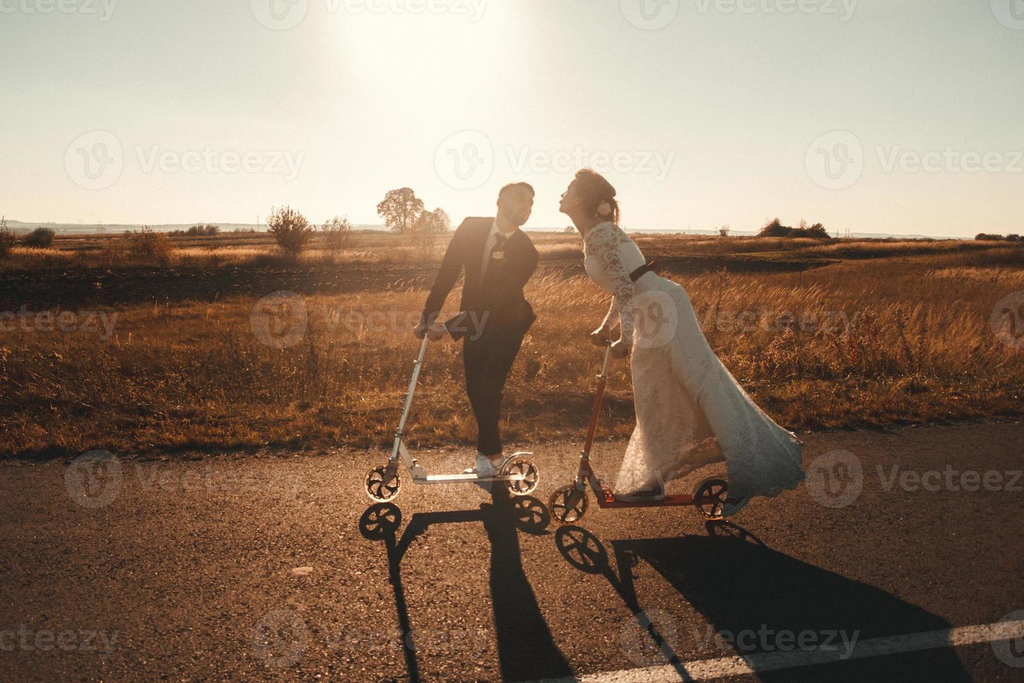 Smiling wedding couple riding a on scooters along the road outside the city at sunset. Place for text or advertising photo