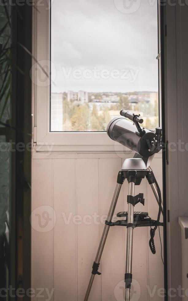 Un gran telescopio gris de pie en el balcón apuntaba hacia el cielo nublado a través de la ventana. sin observador del cielo foto