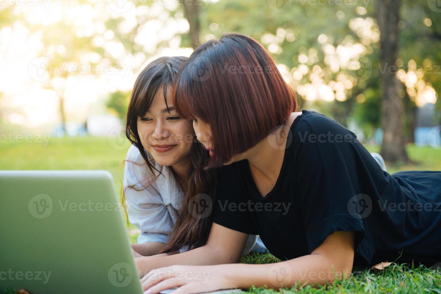 mujeres asiáticas jovenes que yacen en la hierba y que usan la computadora portátil y que escriben. manos de las niñas en el teclado. concepto de aprendizaje a distancia. feliz hipster mujeres asiáticas jóvenes que trabajan en la computadora portátil en el parque. estudiante que estudia al aire libre. foto