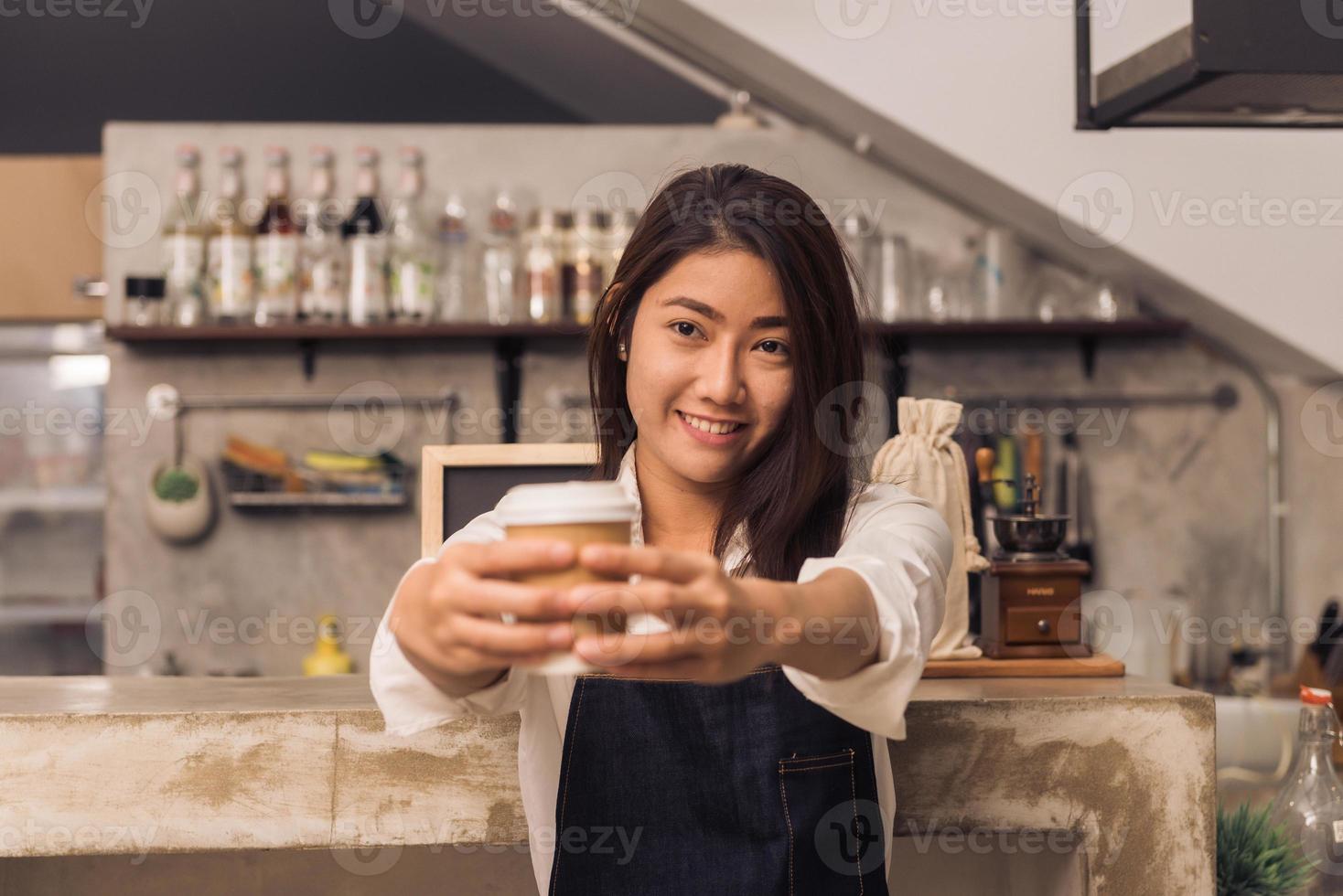 Close up of a young Asian female barista hold a cup of coffee serving to her customer with smile surrounded with bar counter background. Young female barista and her small shop. Food and drink concept photo