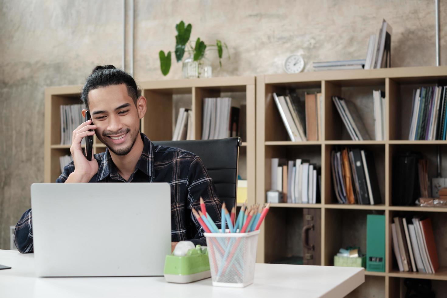 Young male worker of Asian ethnicity, laptop to do creative work, mobile phone communication on desk, bookshelf behind at casual workplace, startup business person, and online e-commerce occupation. photo