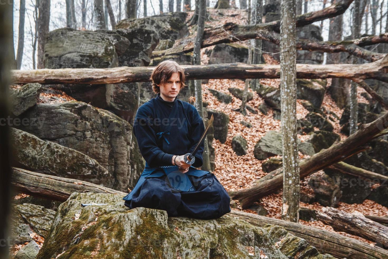 El hombre en kimono negro con una espada medita y se concentra en las rocas y el fondo del bosque foto