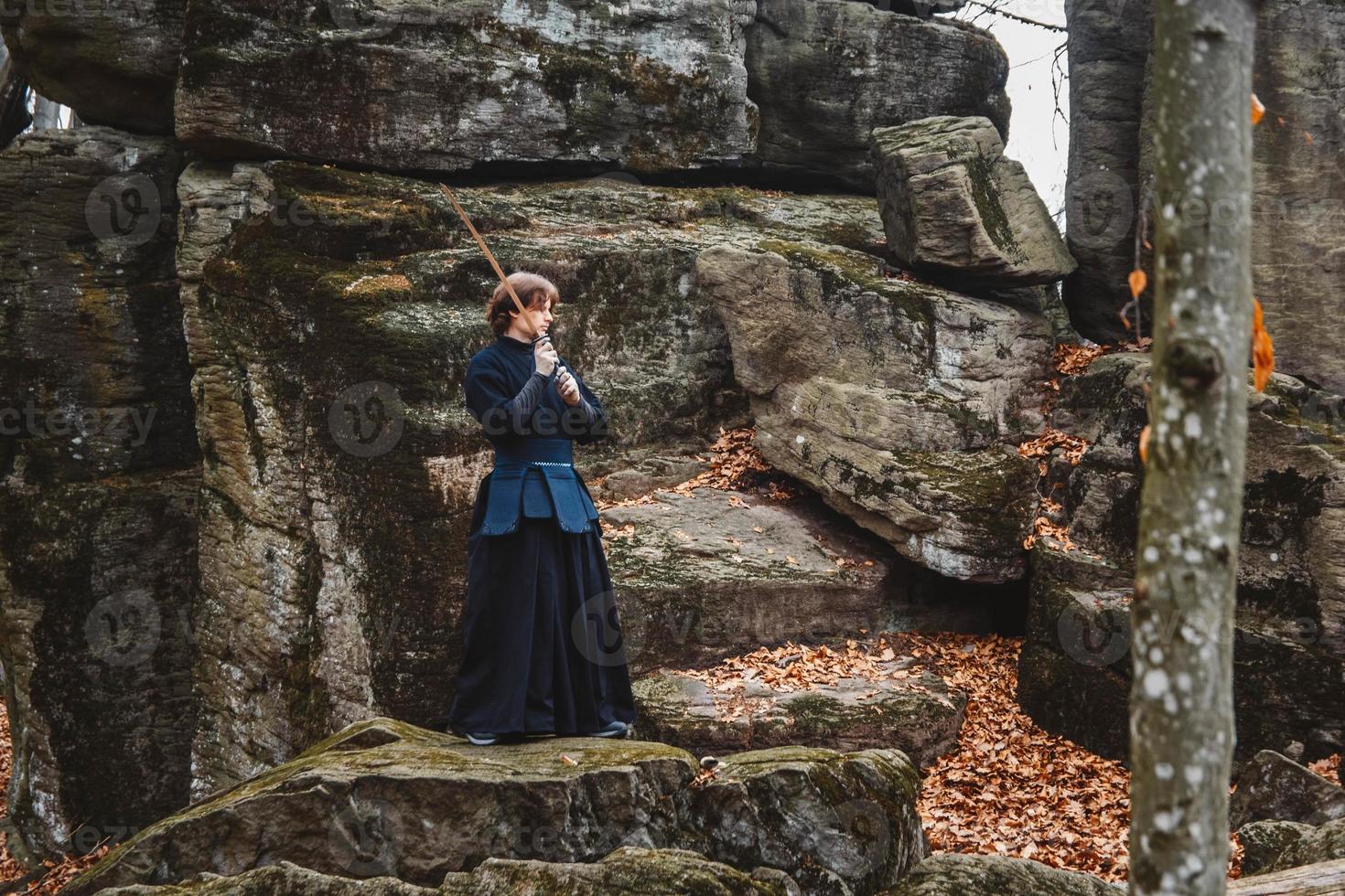 Man in black kimono practicing martial arts with a sword on rocks and forest background photo