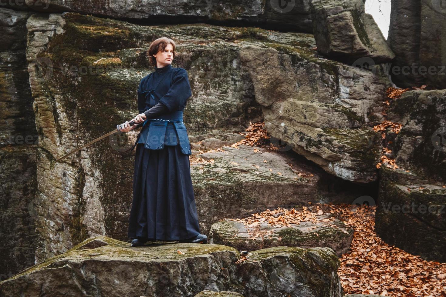 Man in black kimono practicing martial arts with a sword on rocks and forest background photo