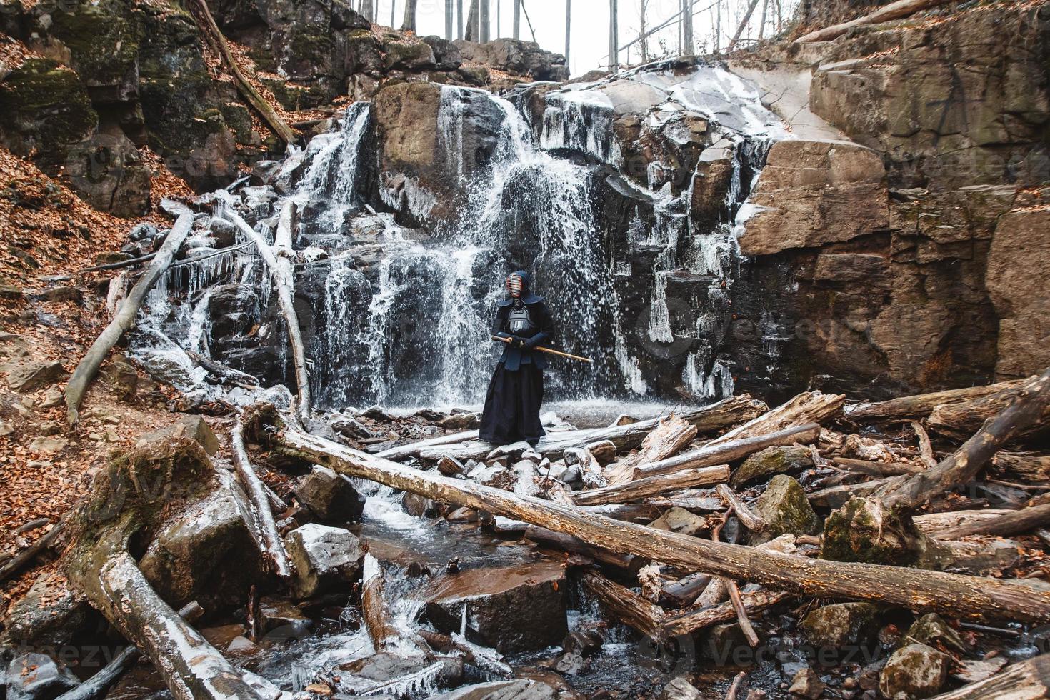 Hombre practicando kendo con espada de bambú en cascada, rocas y fondo de bosque foto