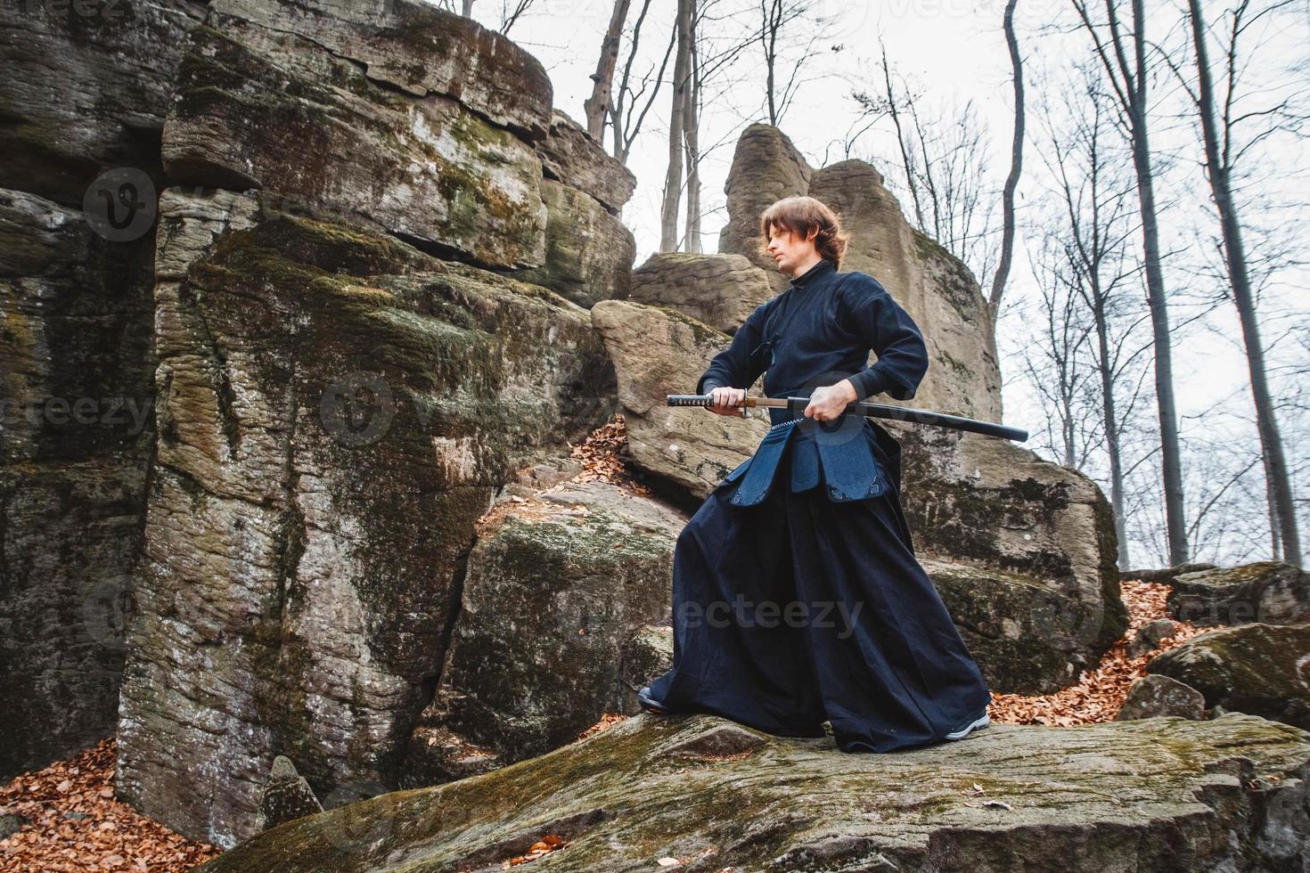 Man in black kimono practicing martial arts with a sword on rocks and forest background photo