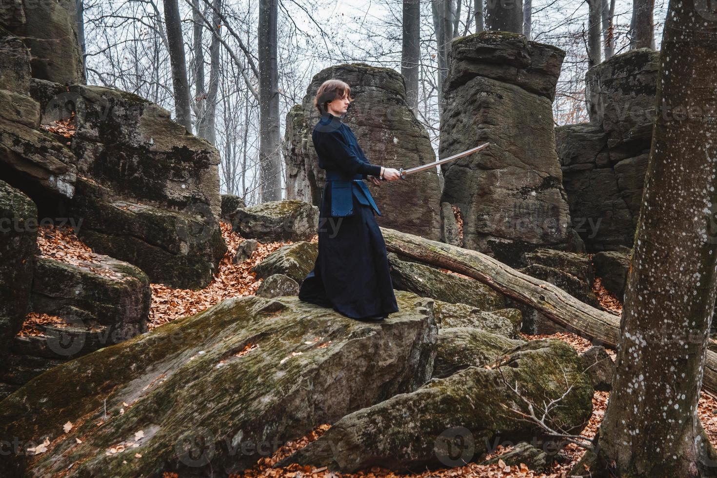 Man in black kimono practicing martial arts with a sword on rocks and forest background photo