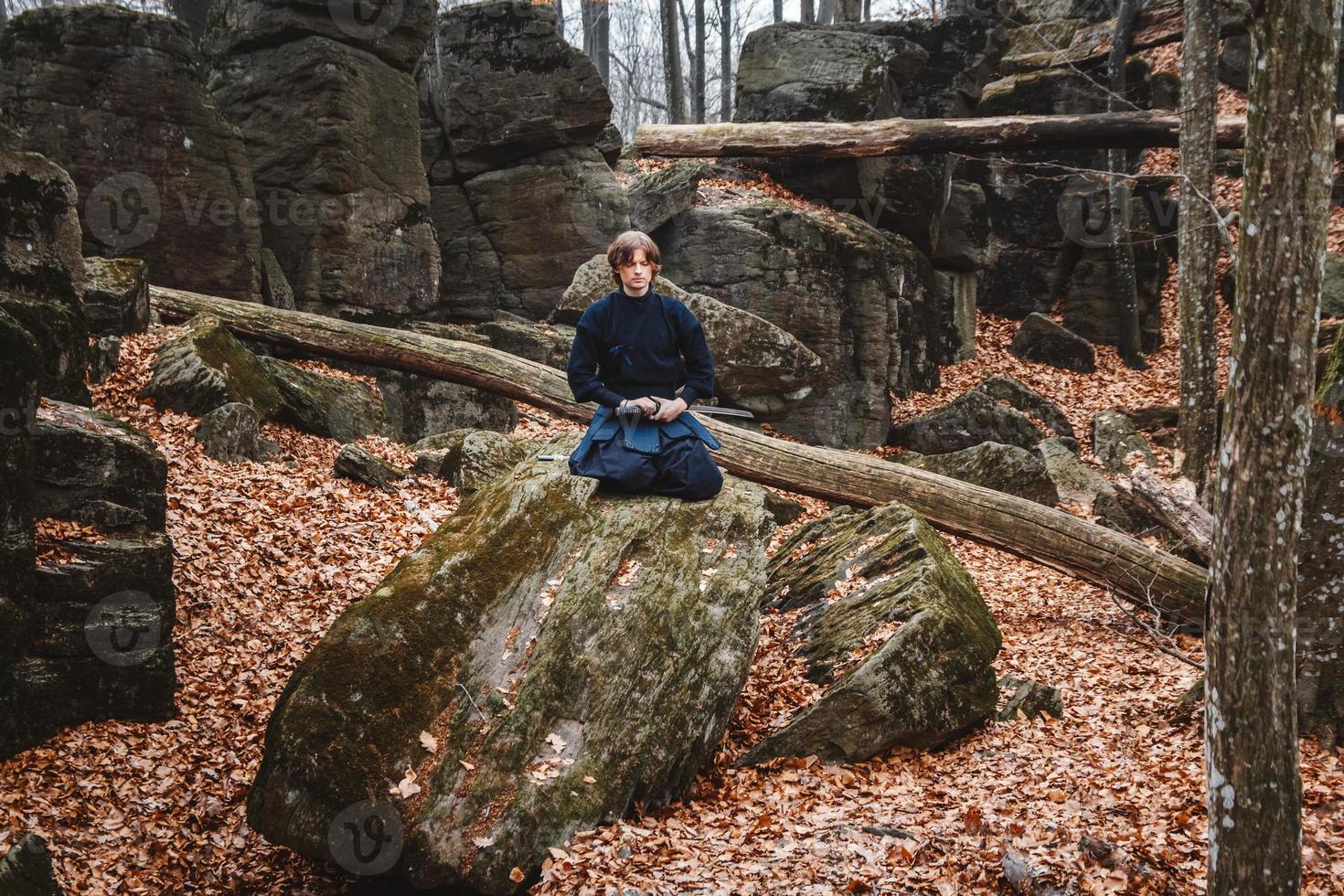 Man in black kimono with a sword meditates and concentrates on rocks and forest background photo