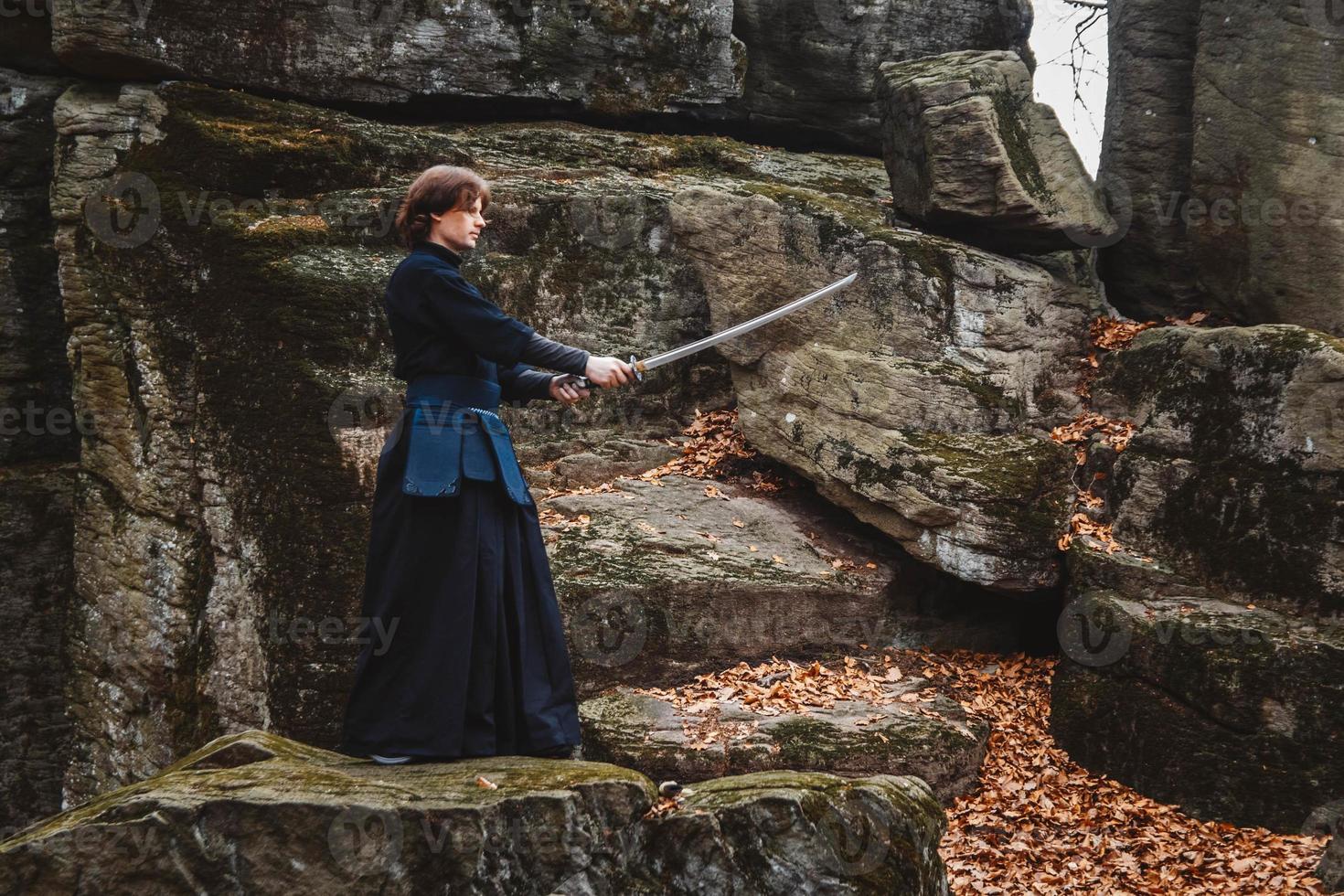 Young man in black kimono practicing martial arts with a sword on rocks and forest background photo