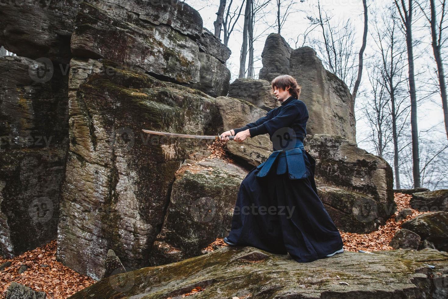 Young man in black kimono practicing martial arts with a sword on rocks and forest background photo