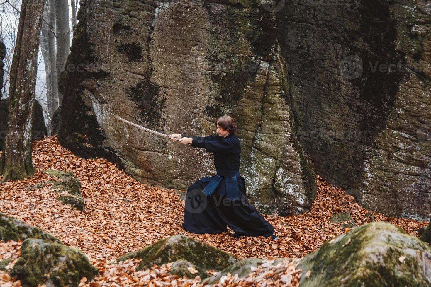 Young man in black kimono practicing martial arts with a sword on rocks and forest background photo