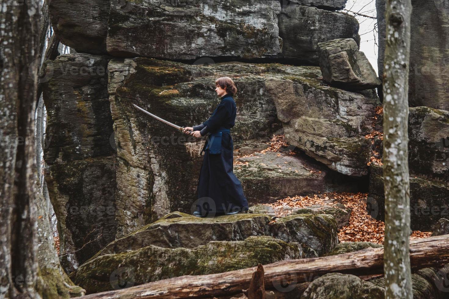 Man in black kimono practicing martial arts with a sword on rocks and forest background photo