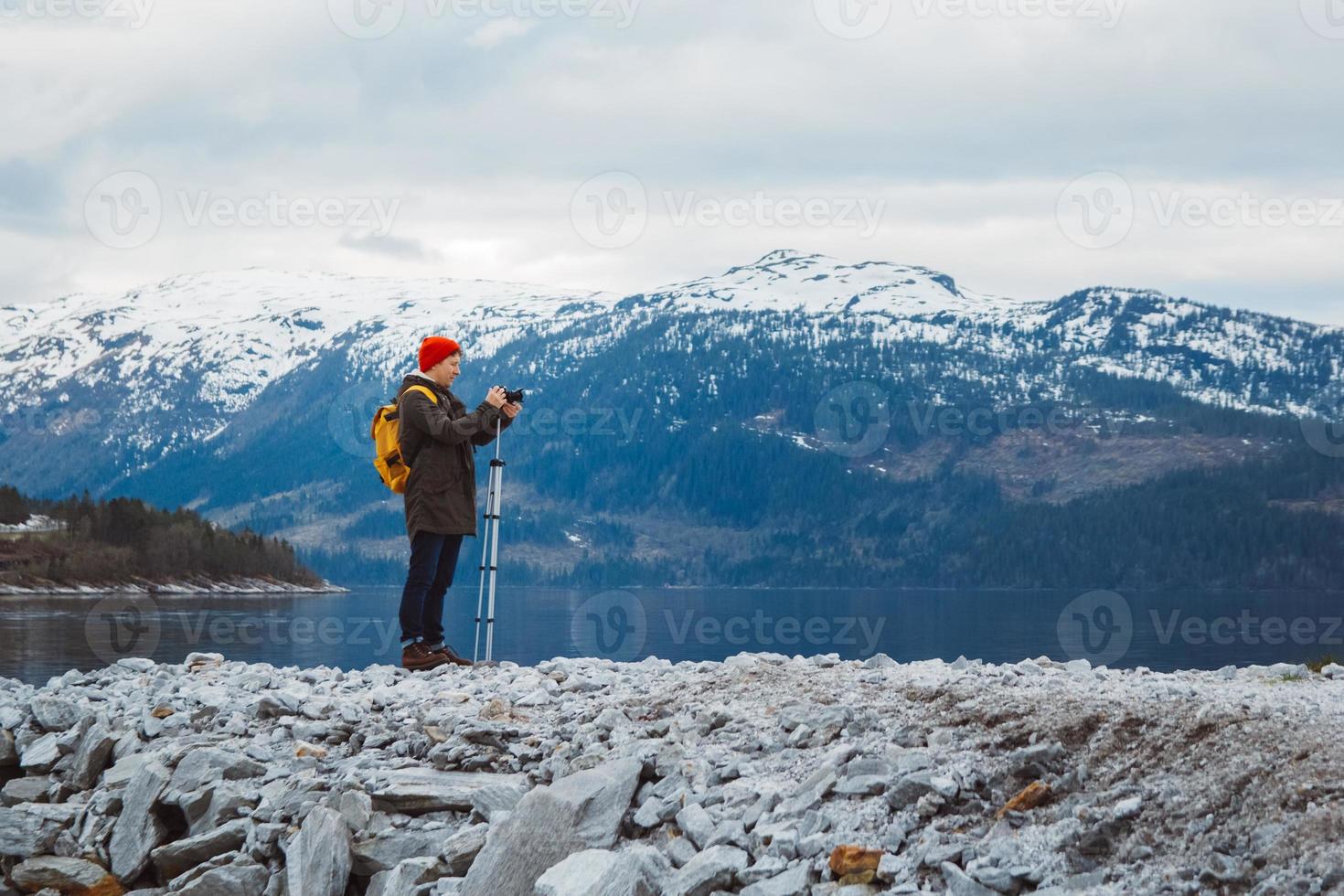Traveler man with camera on tripod standing on background of mountain and a lake makes a photo landscape