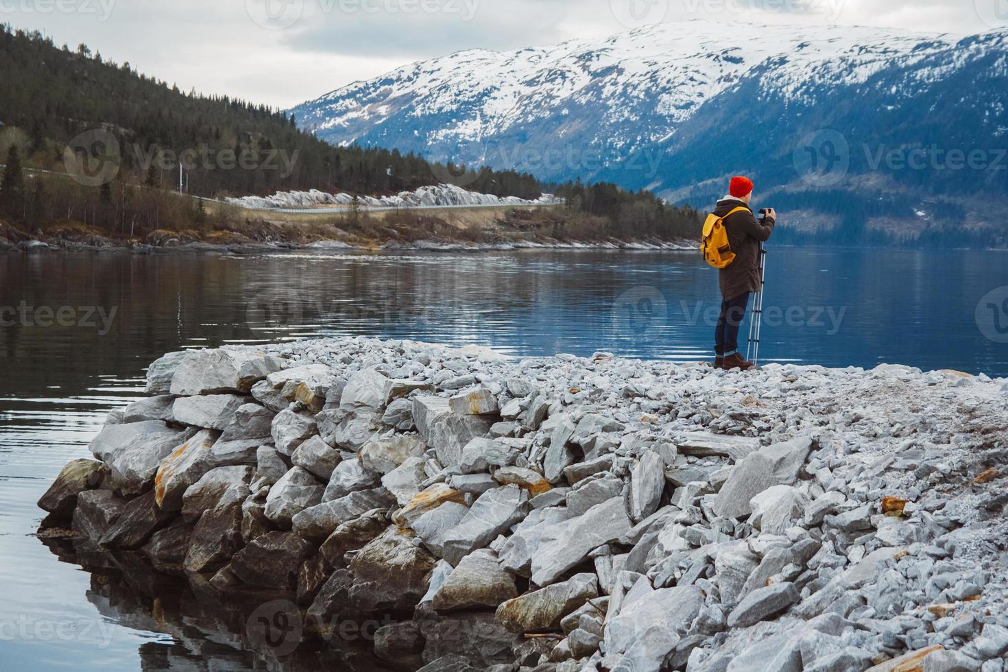 Traveler man with camera on tripod standing on the background of mountain and a lake makes a photo landscape