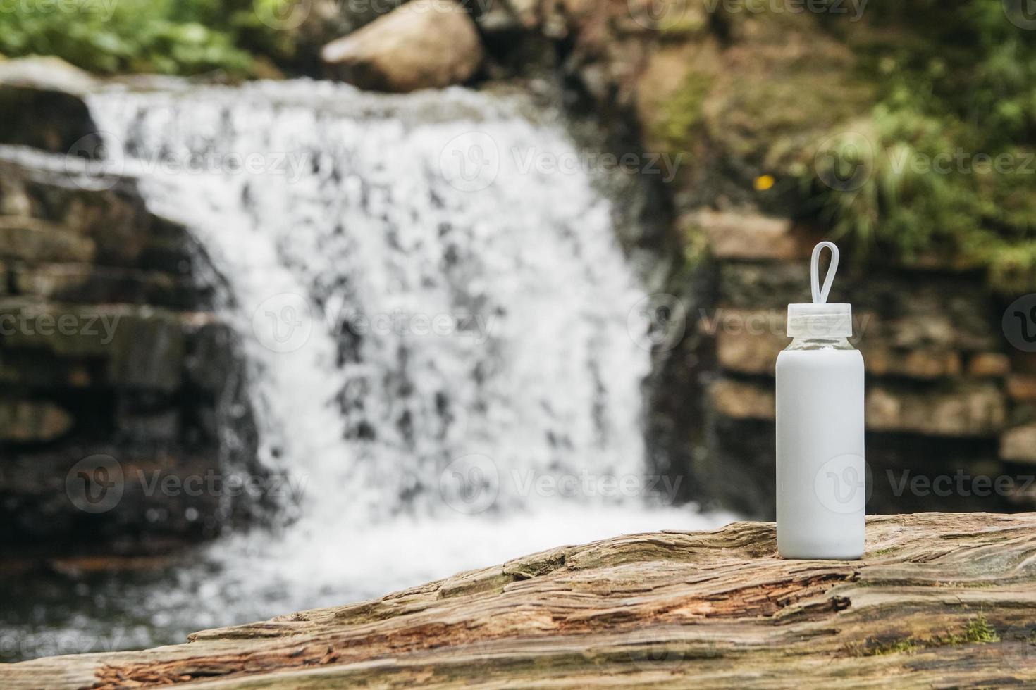 Botella blanca con agua de pie sobre un tronco de madera con el telón de fondo de un río y una cascada foto