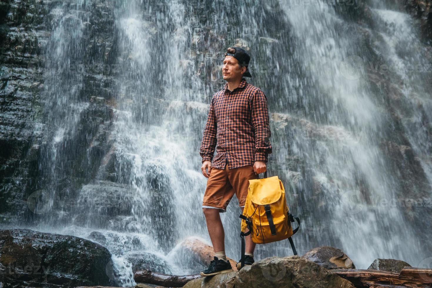 Hombre viajero con una mochila amarilla de pie sobre el fondo de una cascada foto