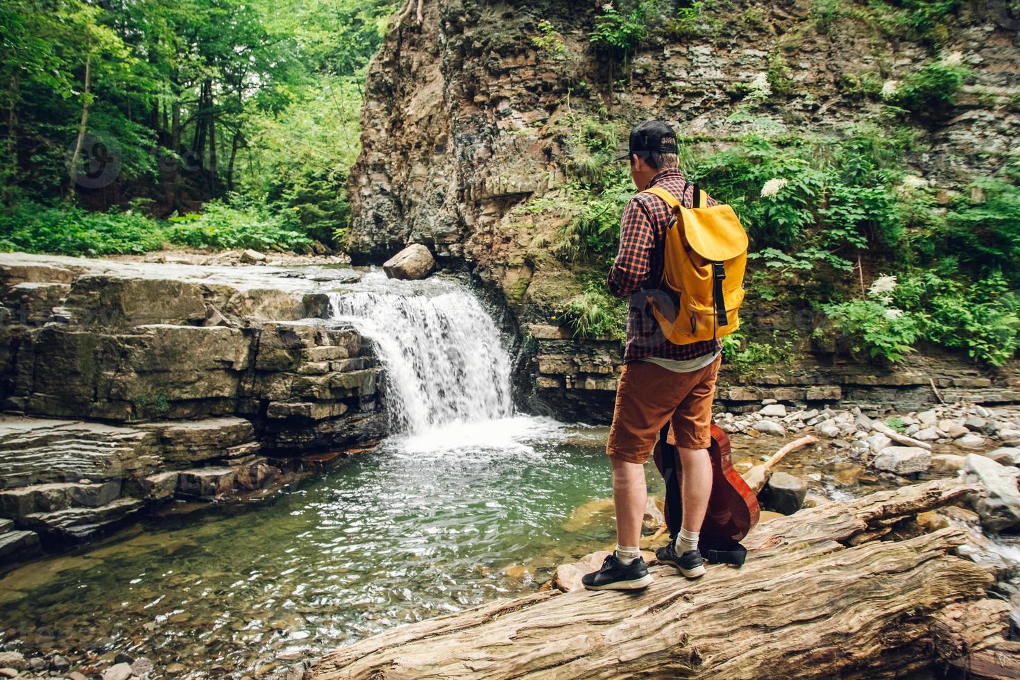 Hombre con una mochila y una guitarra se encuentra en el tronco de un árbol contra una cascada foto