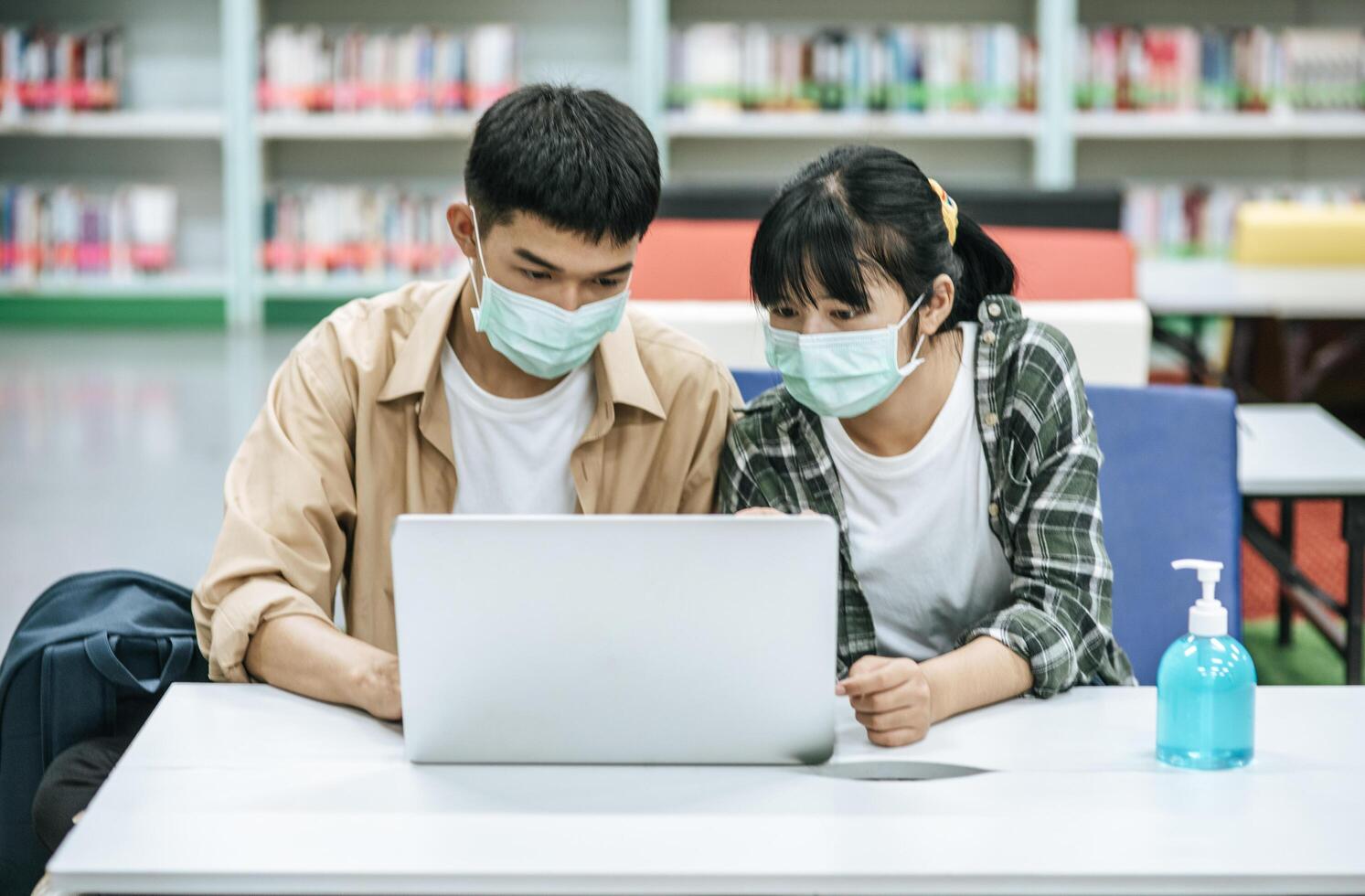 hombres y mujeres usan máscaras y usan una computadora portátil para buscar libros en la biblioteca. foto