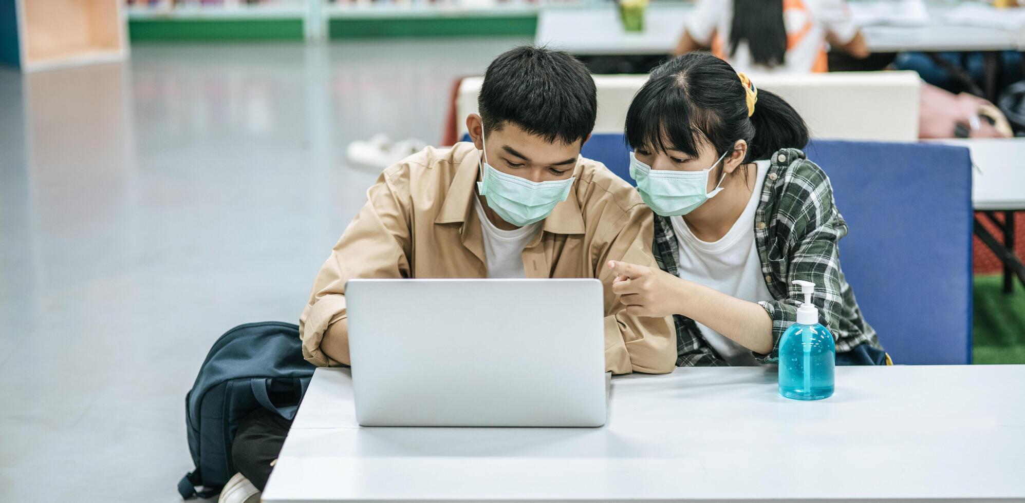 hombres y mujeres usan máscaras y usan una computadora portátil para buscar libros en la biblioteca. foto