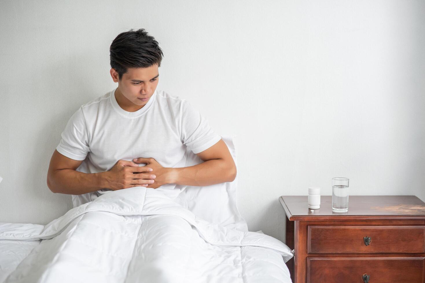 un hombre se sienta en la cama con dolor de estómago y se aprieta el estómago con las manos. foto